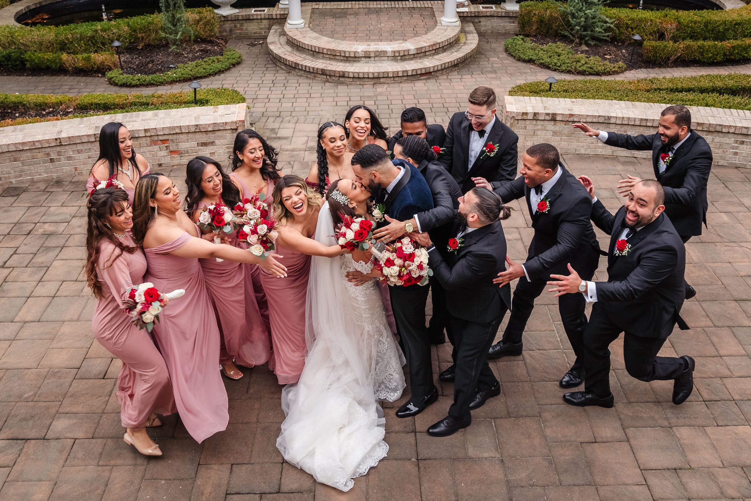 Wedding party in pink bridesmaid dresses and black suits poses playfully on brick patio during outdoor celebration.