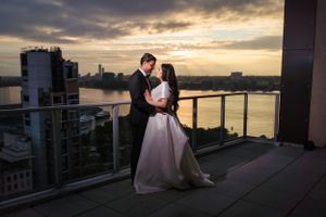 A romantic couple embraces on a high-rise balcony at sunset with dramatic city skyline and water views in the background.