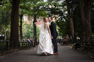A couple in wedding attire dances playfully on a tree-lined path in a park with wooden benches visible in the background.
