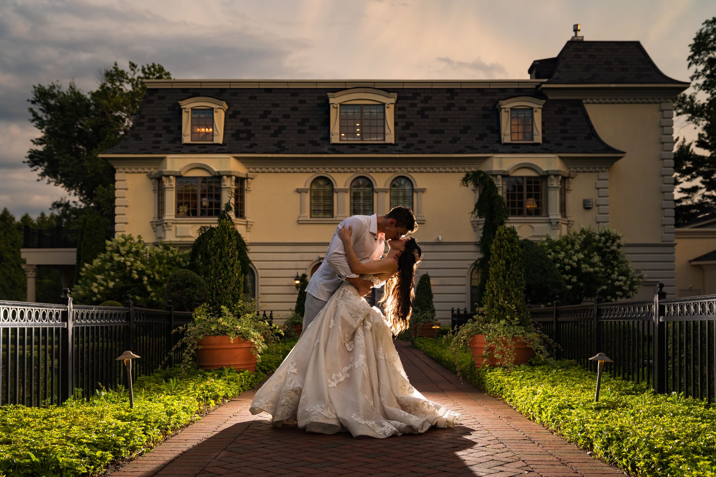 Romantic couple embraces in an elegant dip pose outside a grand manor house at sunset with manicured gardens.