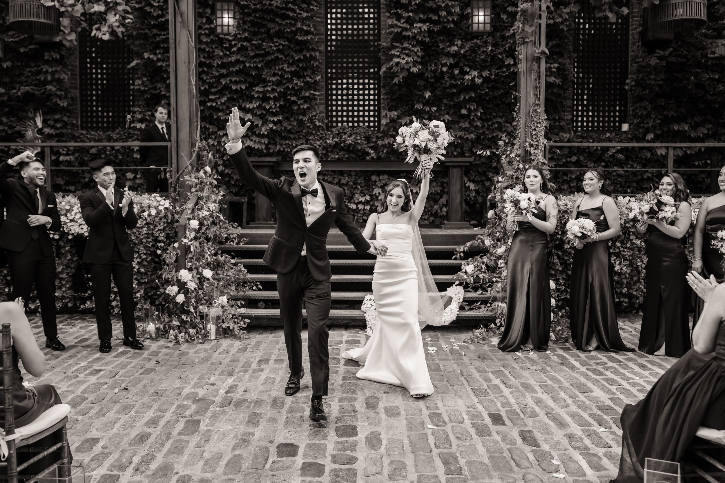 A black and white wedding celebration on brick steps with wedding party members and floral arrangements at an ivy-covered venue.