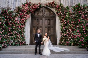 A couple stands in front of an ornate wooden church door framed by a stunning arch of pink and red roses.