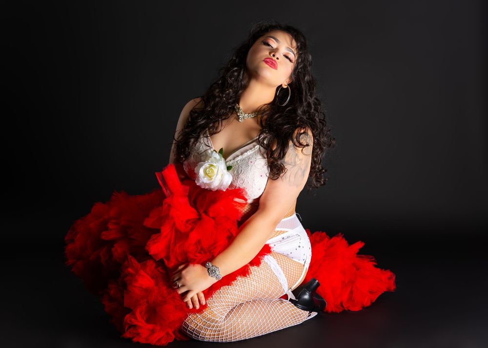Burlesque dancer in white corset and red feather boa poses dramatically on dark stage.
