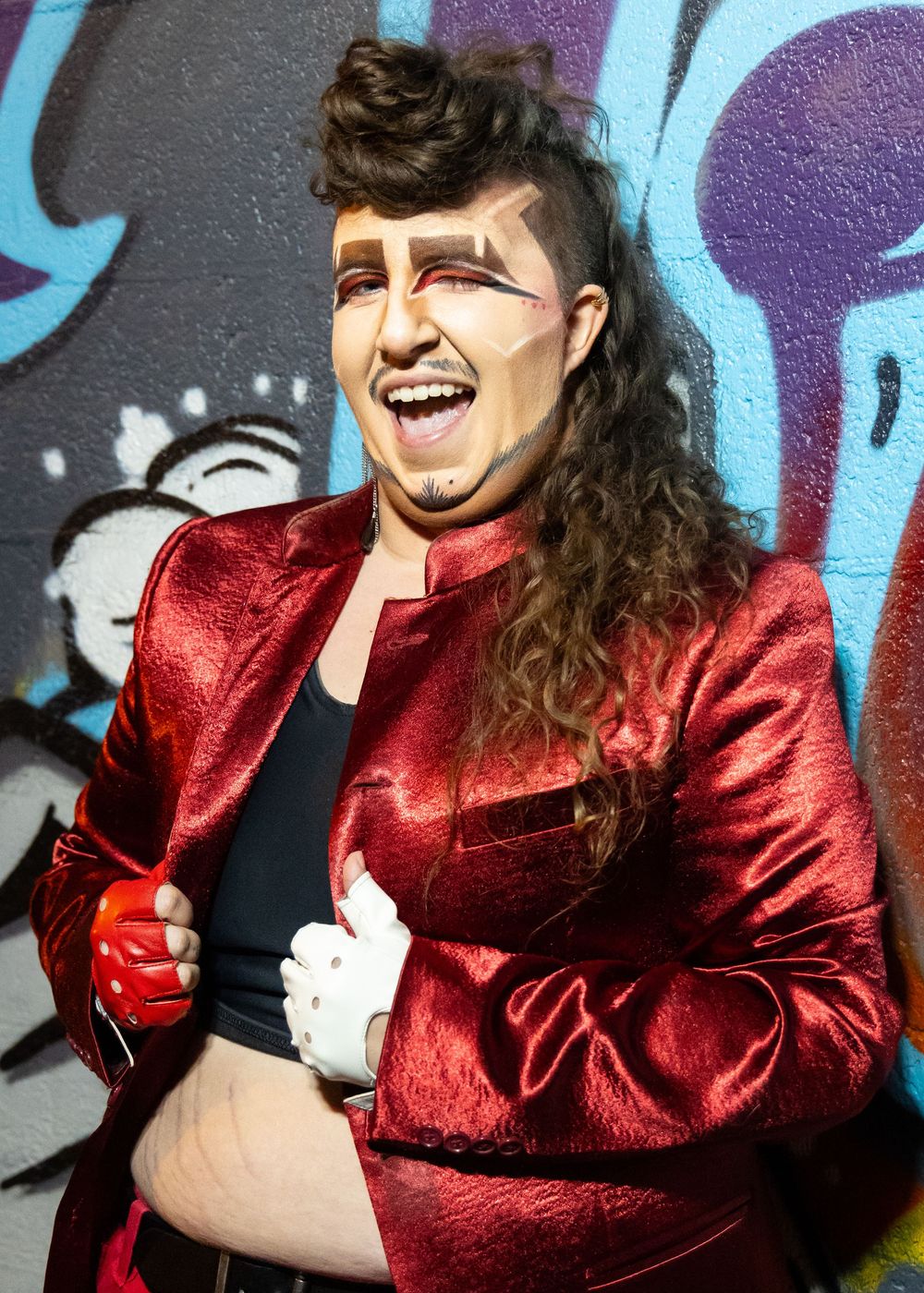 Performer in shiny red jacket laughs joyfully against street art background.