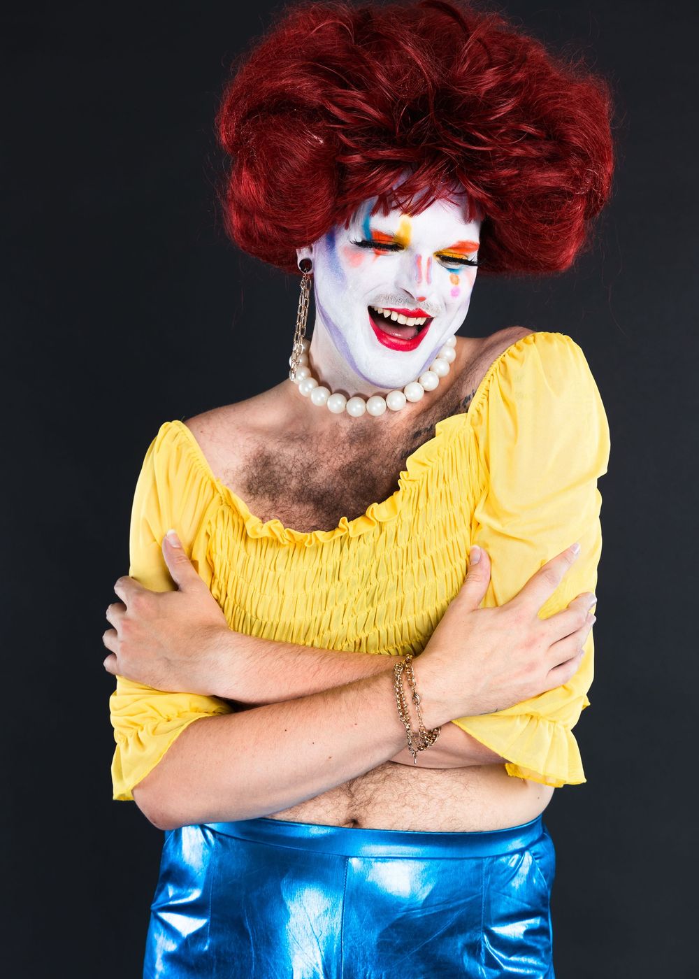 Performer in yellow top and clown makeup poses dramatically on black background.