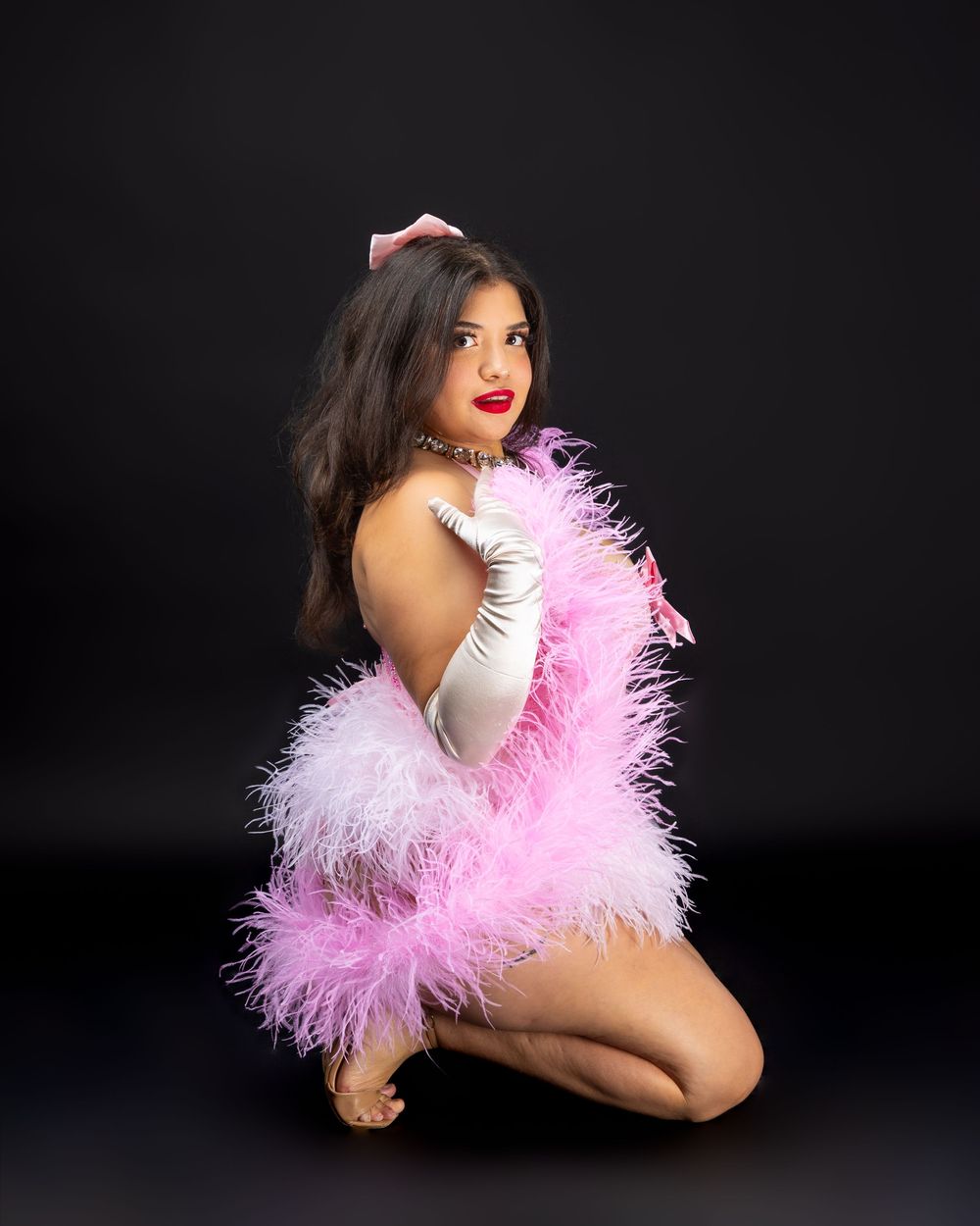 Glamorous burlesque performer poses in pink feather boa and white gloves against dark background.