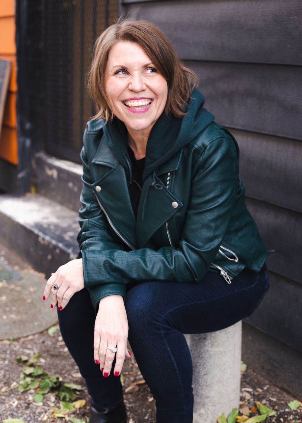 Person in dark leather jacket sitting against black wall outdoors.