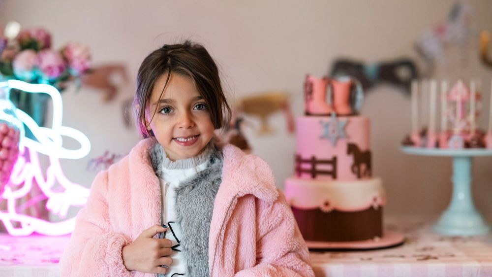 Young person wearing pink coat at birthday party with decorated cakes in background.