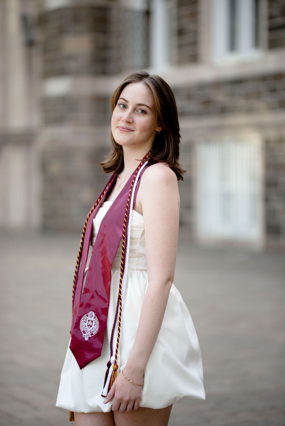 Graduate in a white dress and burgundy graduation stole poses outdoors on campus.