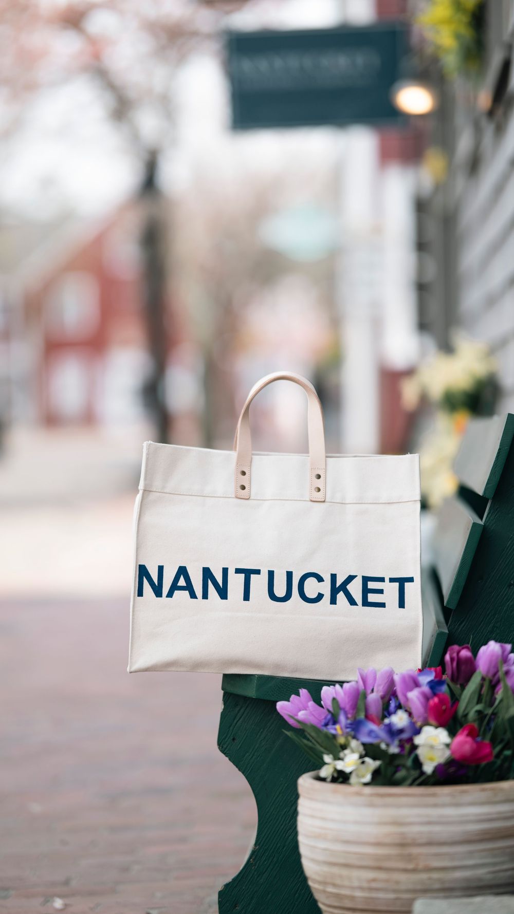 A white canvas tote bag with 'NANTUCKET' text sits beside purple flowers on a cobblestone street.
