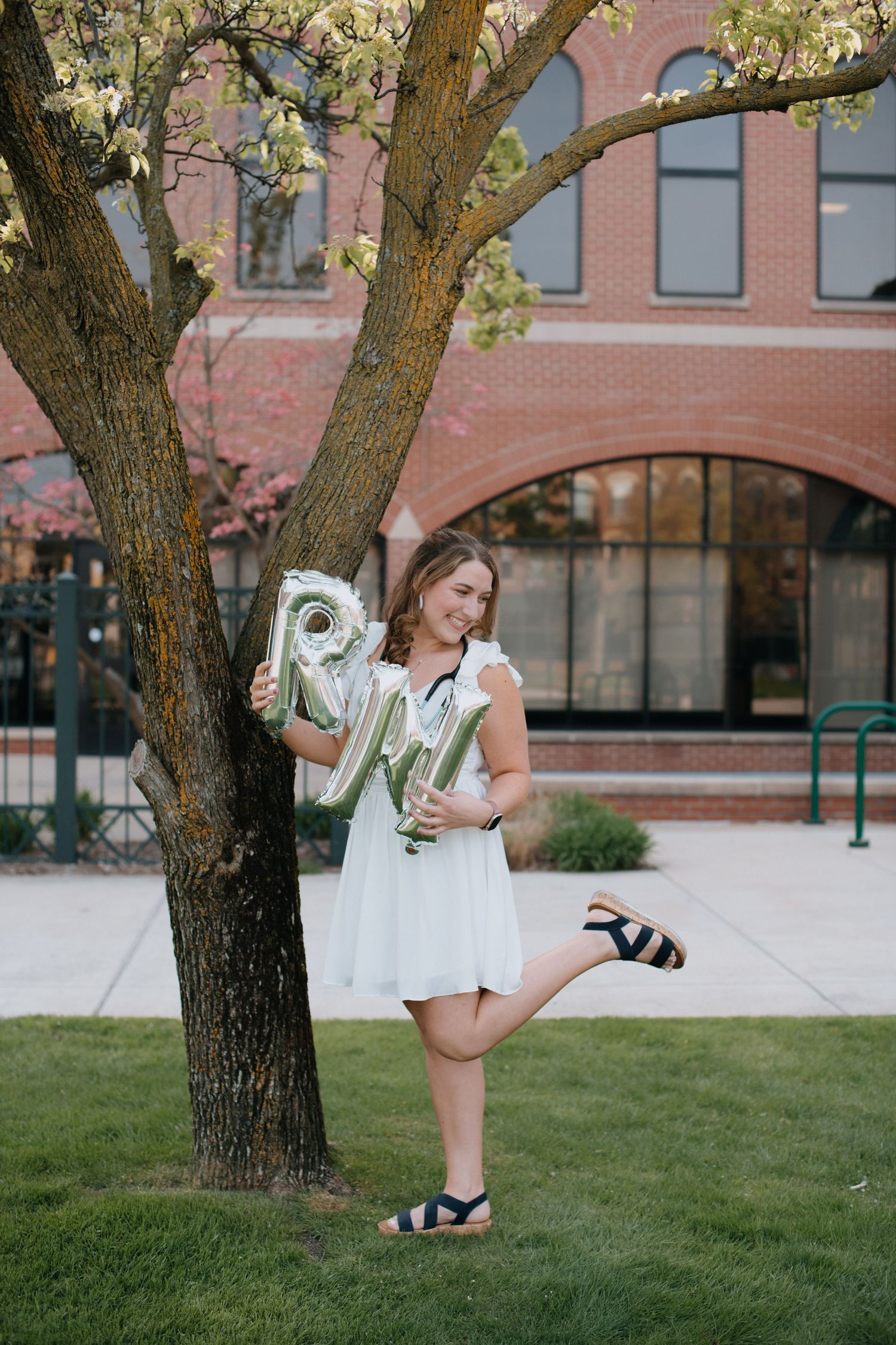 A person in a white dress stands on grass near a brick building, holding letter balloons against a flowering tree.