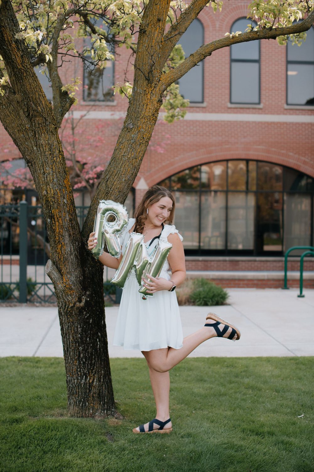 A person in a white dress stands on grass near a brick building, holding letter balloons against a flowering tree.