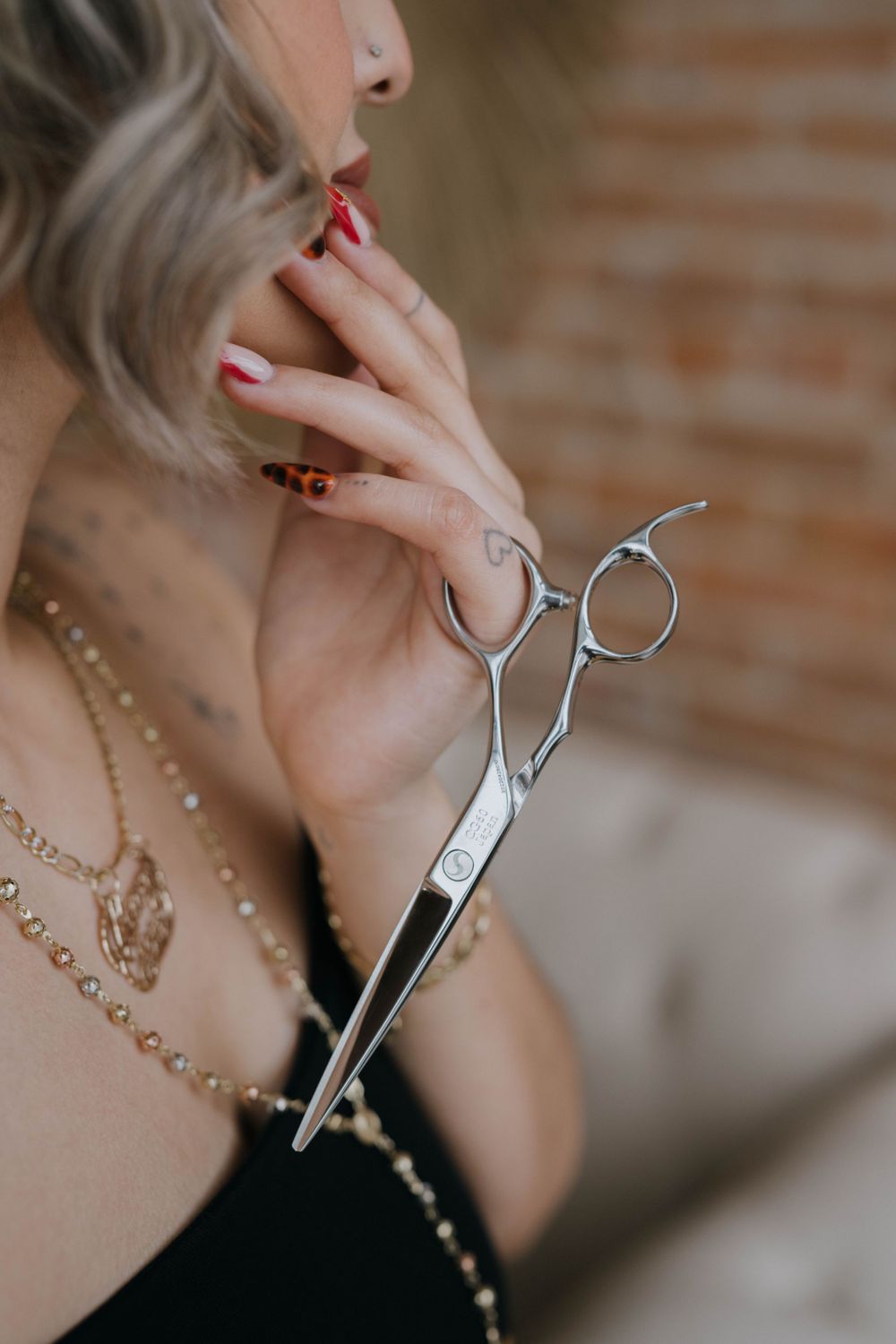 Professional hairstyling scissors held near neck with red manicured nails and delicate gold chain necklace against brick wall.