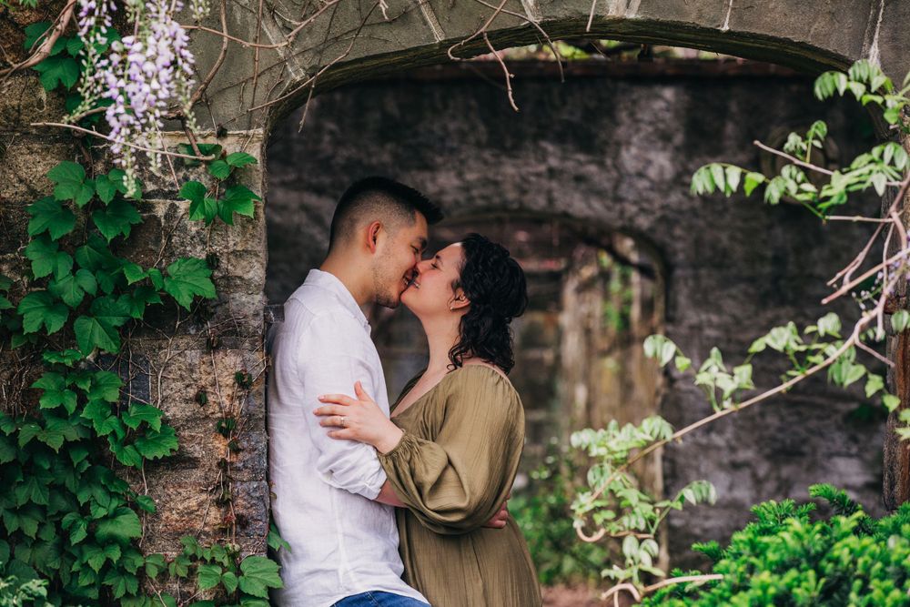 A romantic moment captured under a stone archway surrounded by blooming wisteria and lush greenery.