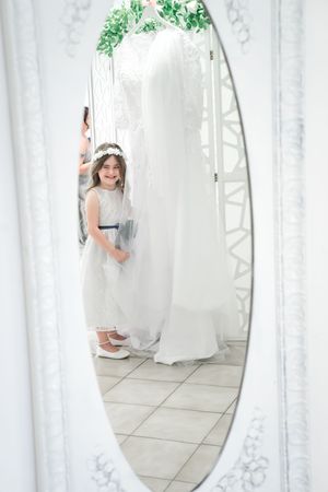 Mirror reflection in a white room shows a small figure in a flowing white dress with greenery decorating the top of the mirror.