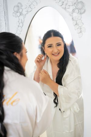 Bride in white robe smiling while putting on earrings in front of ornate mirror.