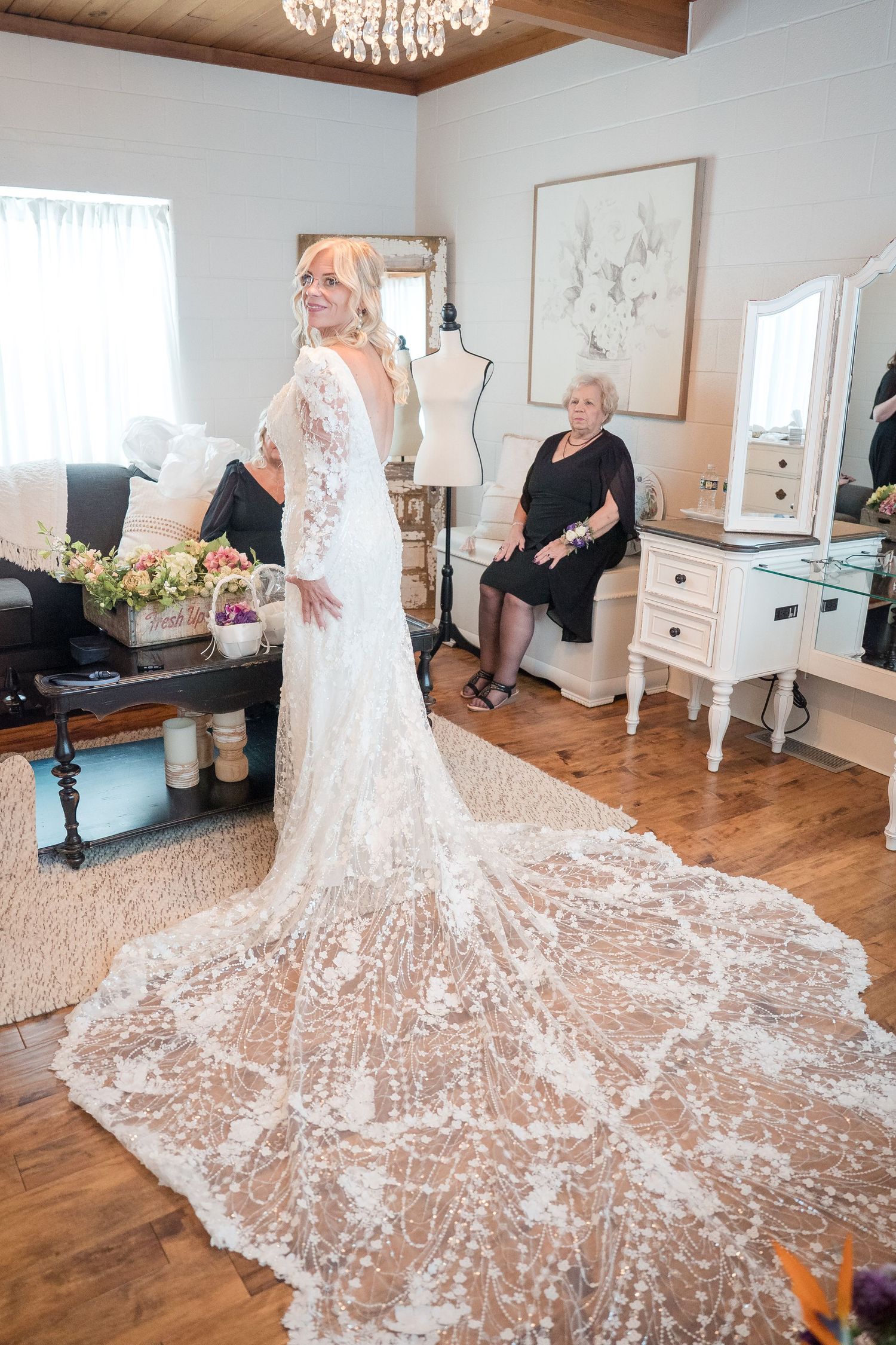 Stunning lace wedding dress with dramatic cathedral train displayed on hardwood floor beneath crystal chandelier.