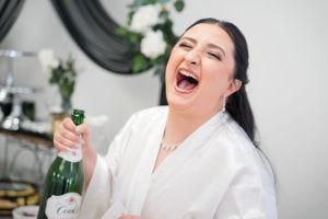 Joyful woman in white robe laughing while holding champagne bottle during wedding preparations.