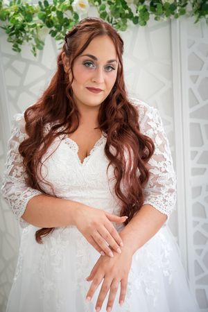 A bride in a white lace wedding dress poses gracefully in front of a floral backdrop for a series of bridal portraits.