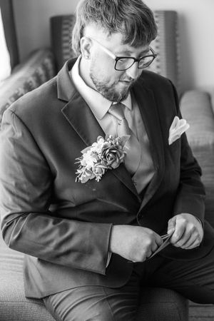 Black and white portrait of groom in suit and glasses wearing floral boutonniere sitting on couch.