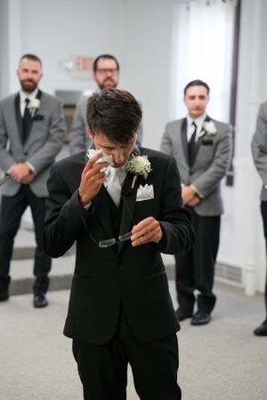 Emotional groom in black suit wipes tears during ceremony while groomsmen in grey suits watch in background.
