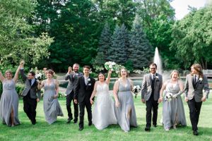 Wedding party in grey and black attire celebrating and jumping outdoors with evergreen trees in background.