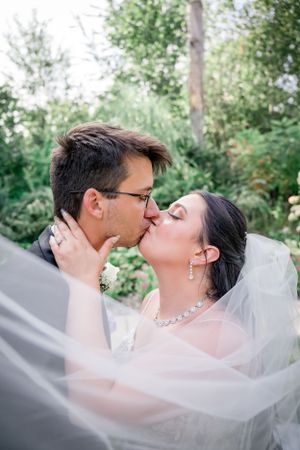 Intimate wedding kiss captured through flowing white veil with natural garden backdrop.