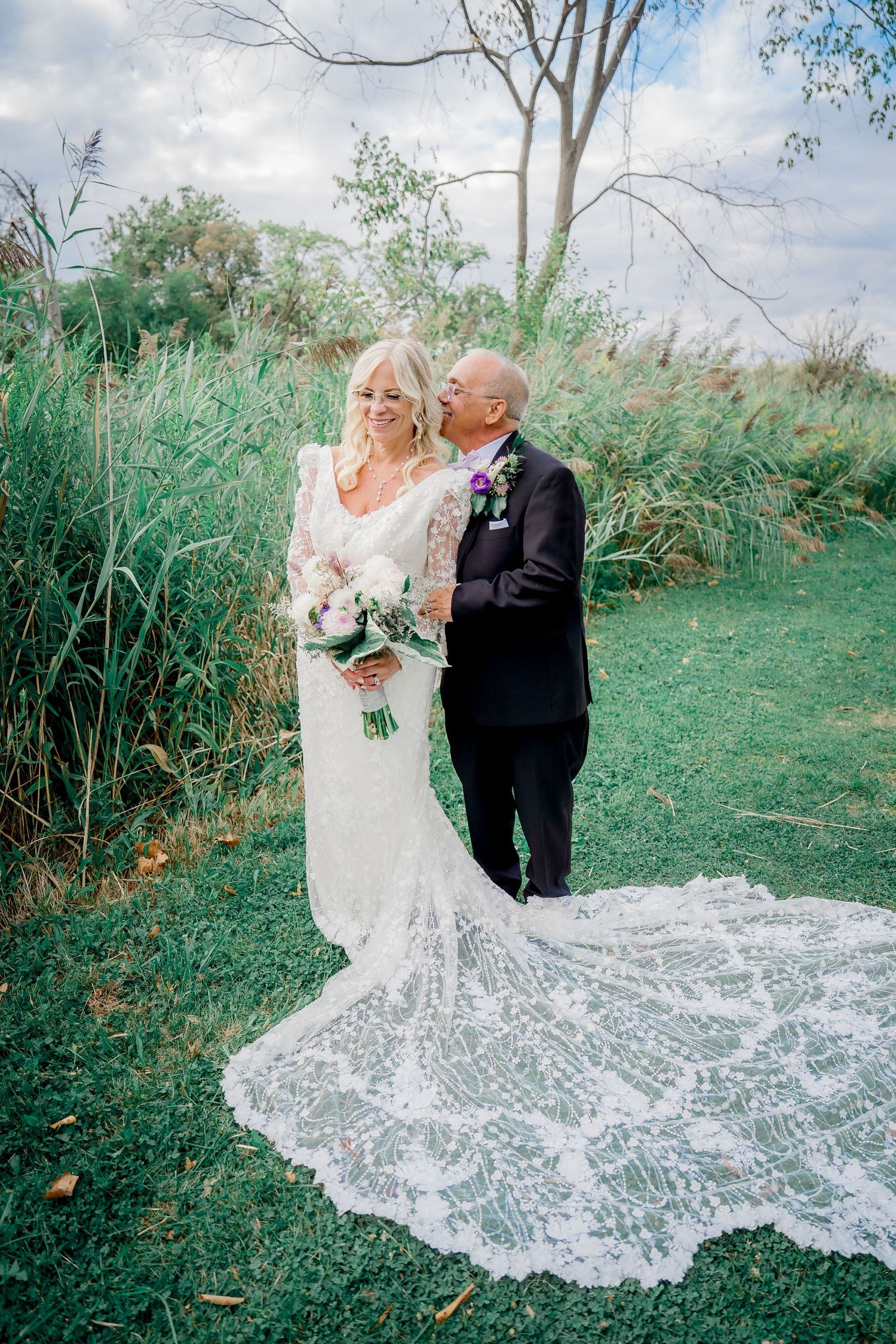 A couple in formal wedding attire pose together outdoors with a flowing lace train spread across green grass.