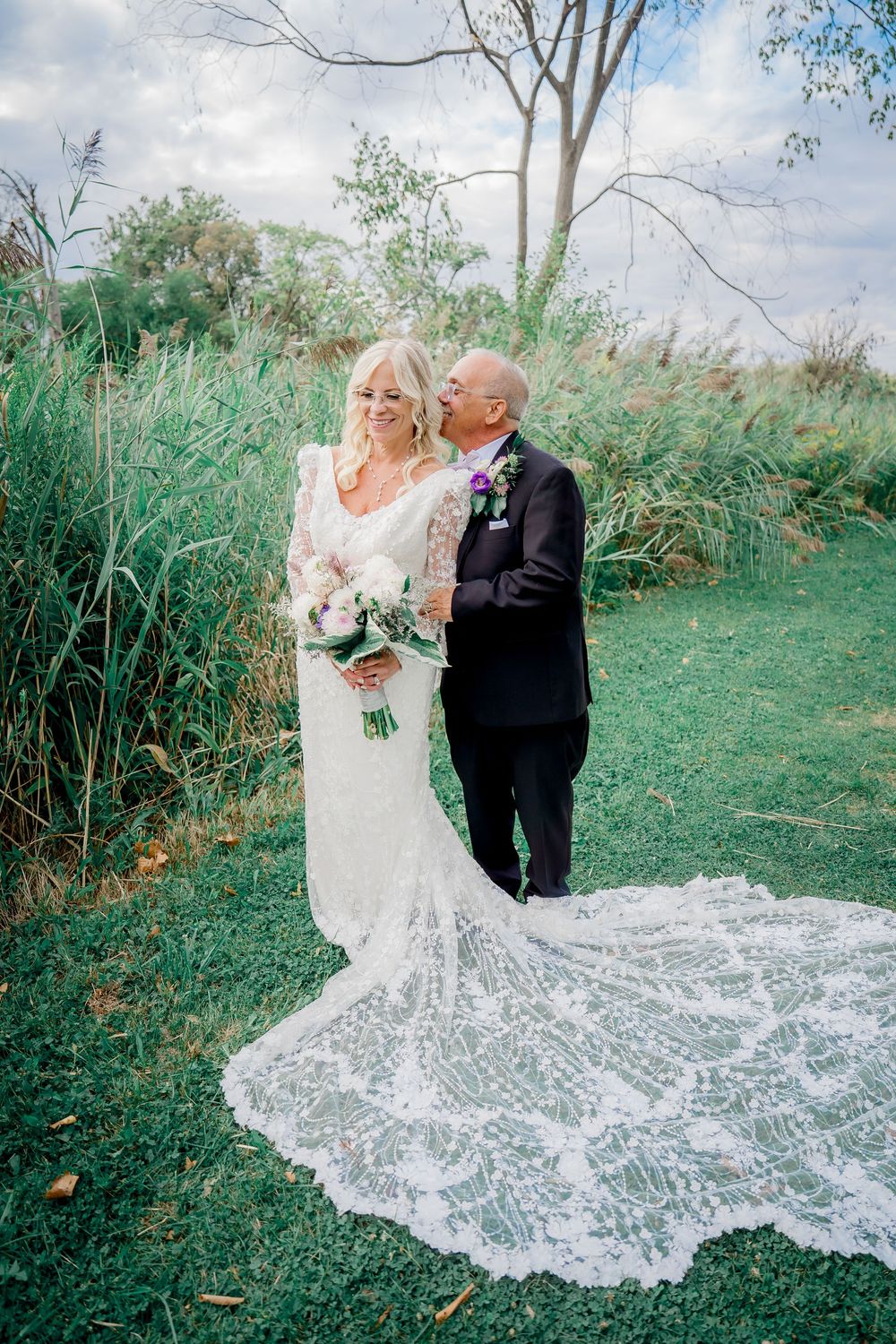 A couple in formal wedding attire pose together outdoors with a flowing lace train spread across green grass.