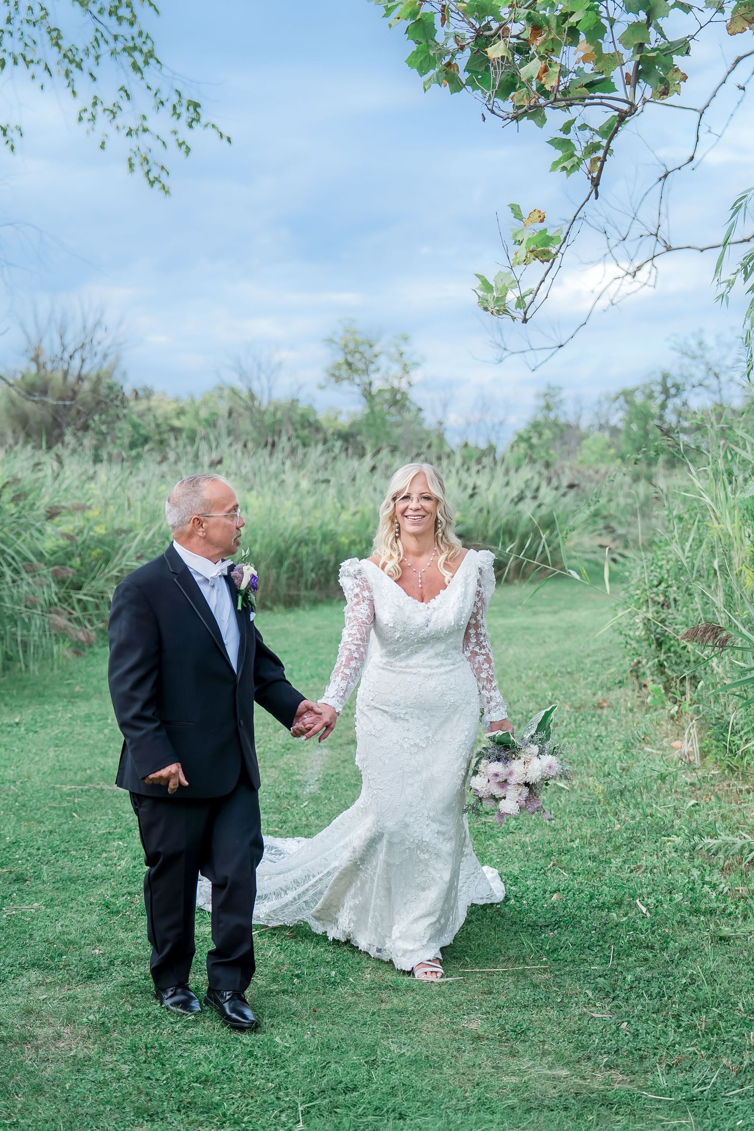 A couple in formal wedding attire walks hand in hand on a grassy path with natural greenery and blue sky in the background.