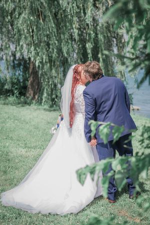 Newlywed couple in elegant wedding attire share romantic moments under cascading willow tree branches in a garden setting.