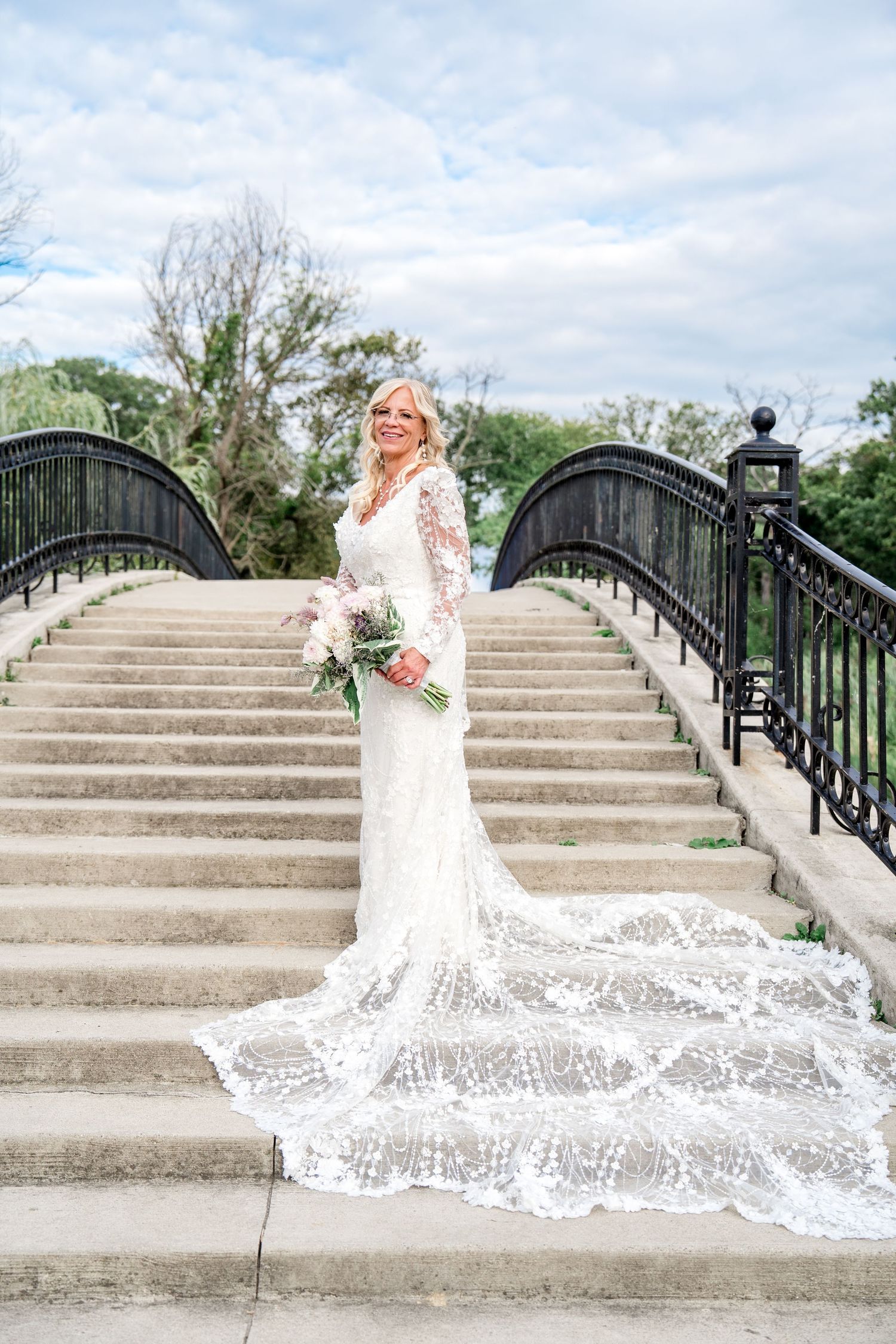 A bride in a flowing lace wedding dress poses elegantly on concrete steps with ornate black railings.