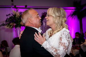 A couple shares an intimate dance moment at a wedding reception with purple lighting and white decor in the background.