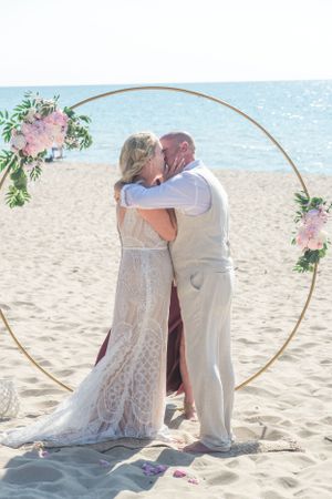 A romantic beach wedding ceremony with a circular arch decorated with pink flowers as the couple shares their first kiss.