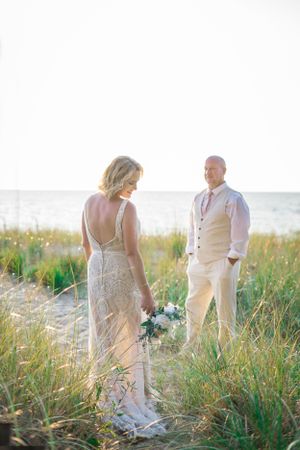 A couple stands in beach grass during golden hour wearing elegant beige and white wedding attire.