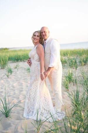 A beachside wedding portrait featuring a couple in elegant white and beige attire among dune grass.