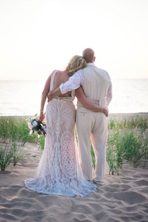 A couple embracing while looking at the ocean, wearing elegant wedding attire on a sandy beach.
