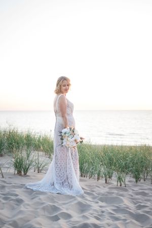 A bride in a lace wedding dress holds a bouquet while standing on a beach at sunset.
