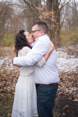 A couple in white and blue outfits share a romantic winter moment in a snowy forest setting.
