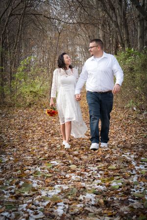 A couple walks together through fallen autumn leaves in a wooded area during a romantic outdoor photoshoot.