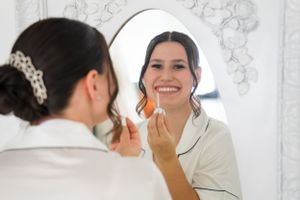 Woman applying makeup while looking in mirror wearing silk pajamas.
