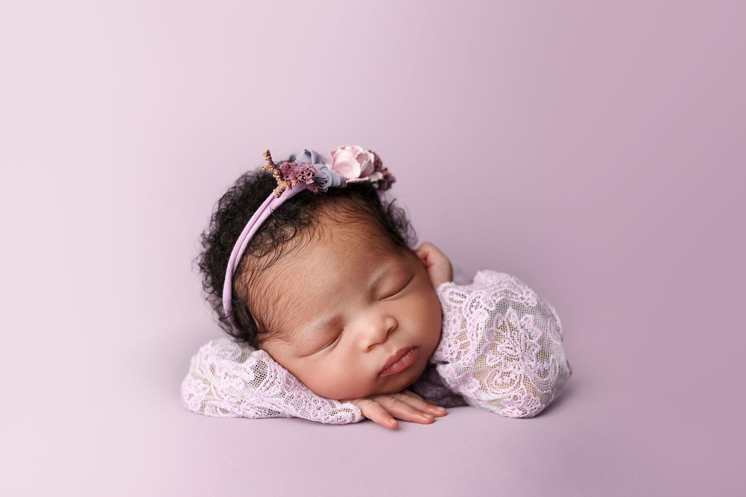 Sleeping newborn wearing floral headband and pink dress on rosy background.