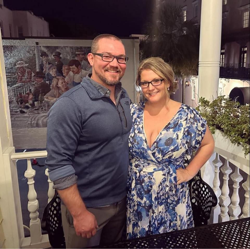 Smiling couple posing together on evening porch, man in blue shirt and woman in floral blue dress.