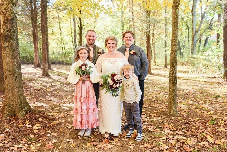 Wedding family portrait with bride, groom and three children standing on leaf-covered forest path.