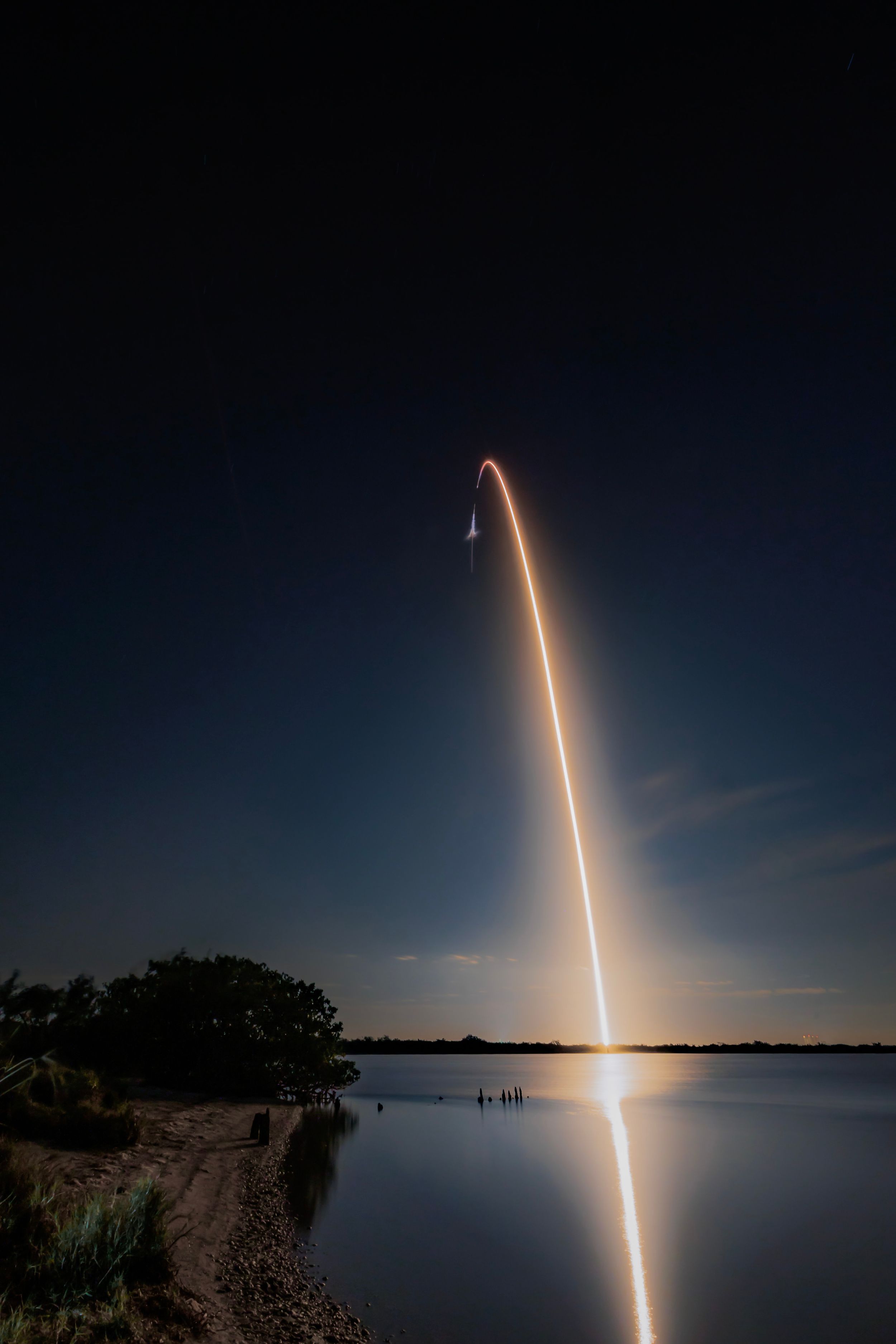 Rocket launch trail reflects in calm water at night as glowing streak arcs across dark sky over shoreline.