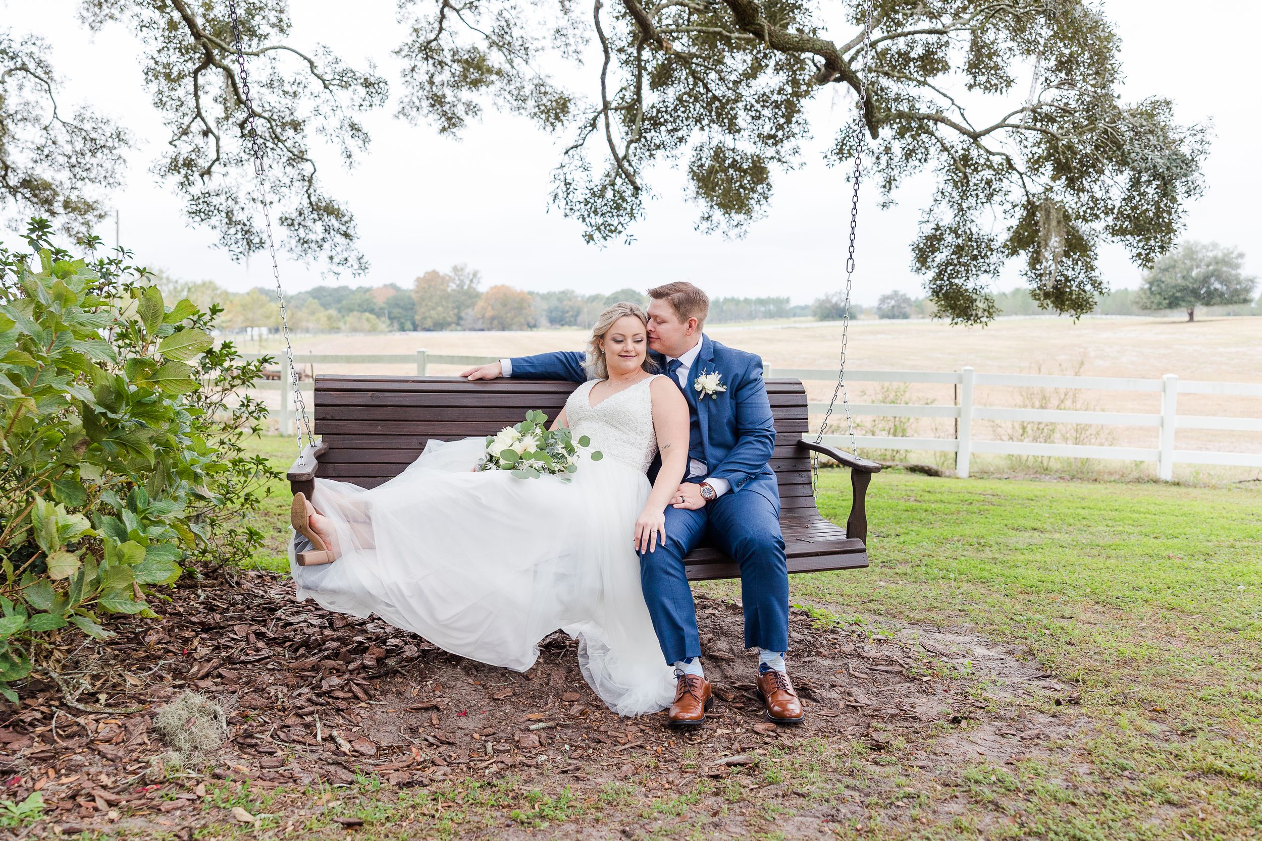 Couple sitting together on a rustic wooden bench under trees at an outdoor venue during a romantic moment.