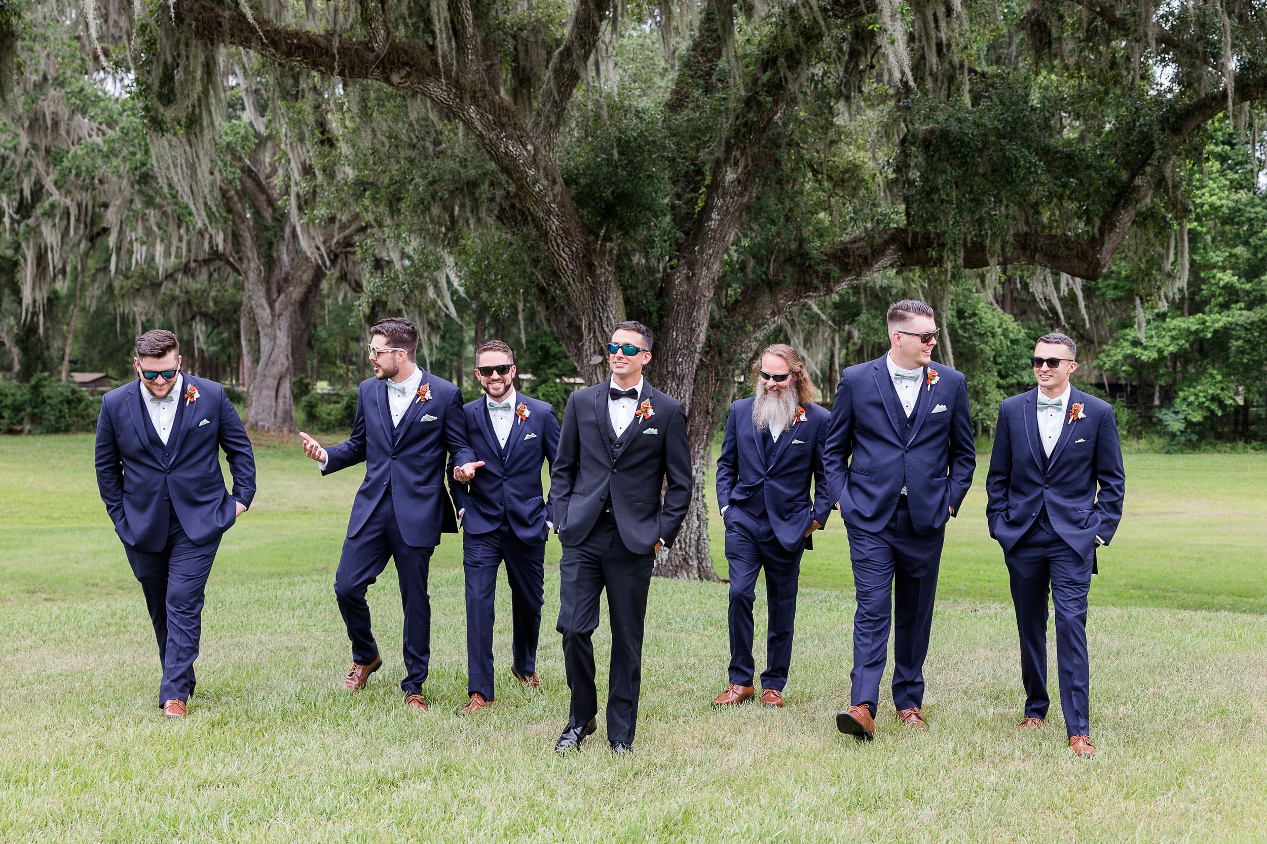 Wedding party in navy suits walks together across a lush green lawn on a sunny day.