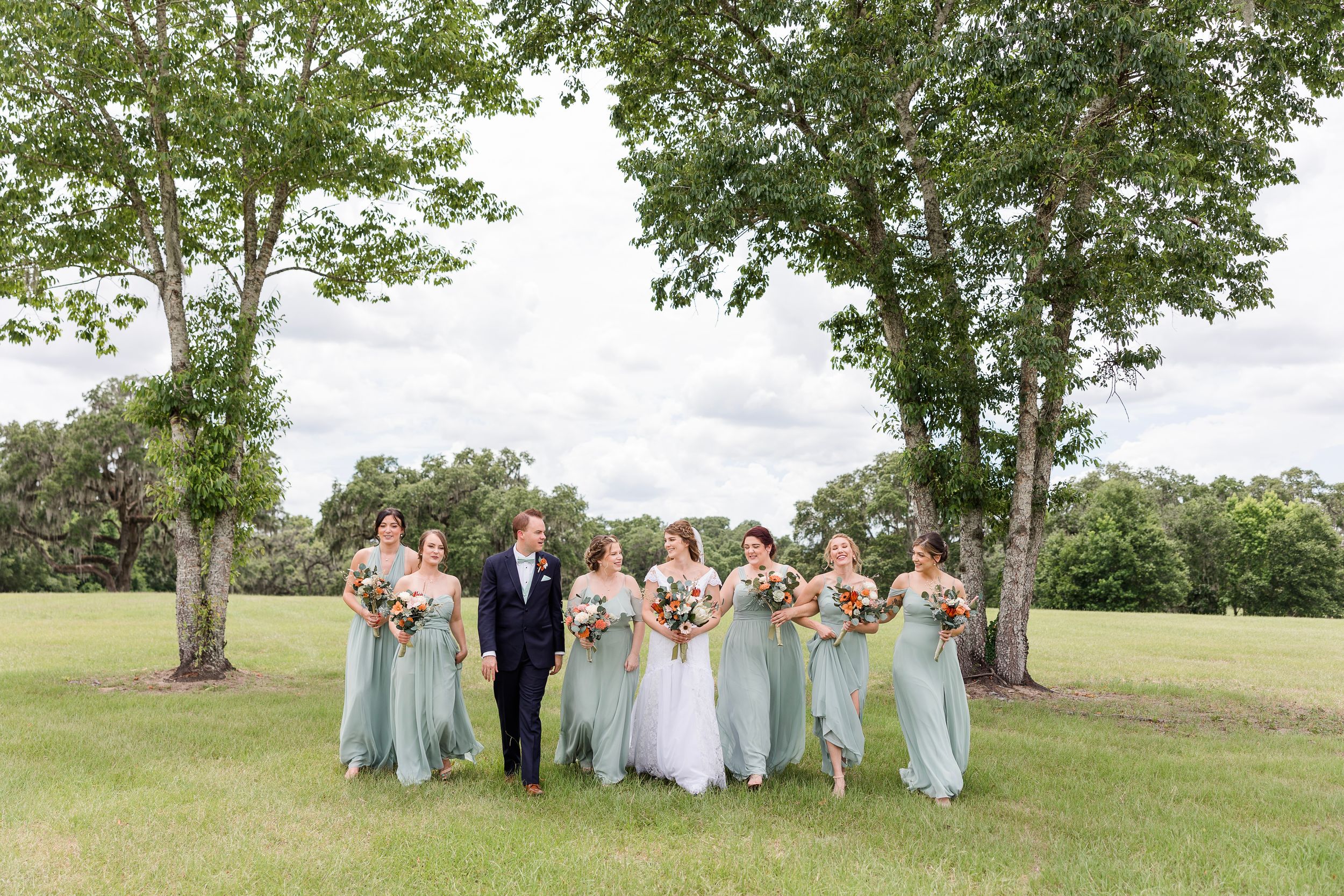 Wedding party in light blue dresses poses together on a grassy hillside during an outdoor summer celebration.