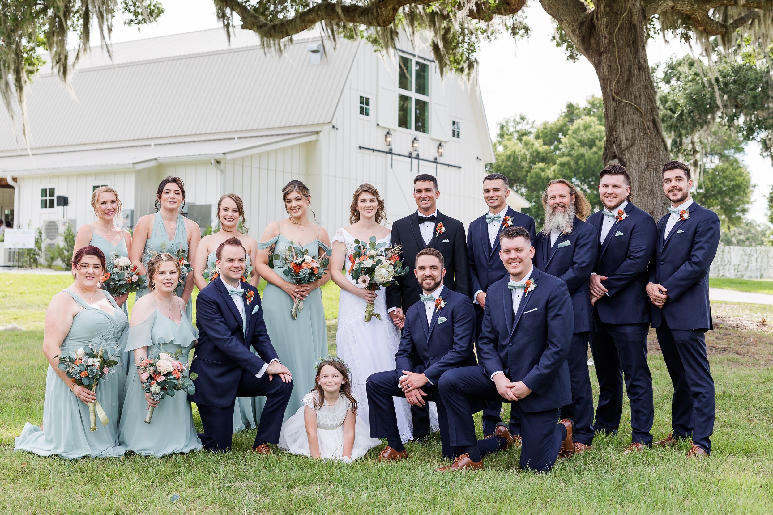 Wedding party in mint green dresses and navy suits poses together under a large oak tree at outdoor venue.