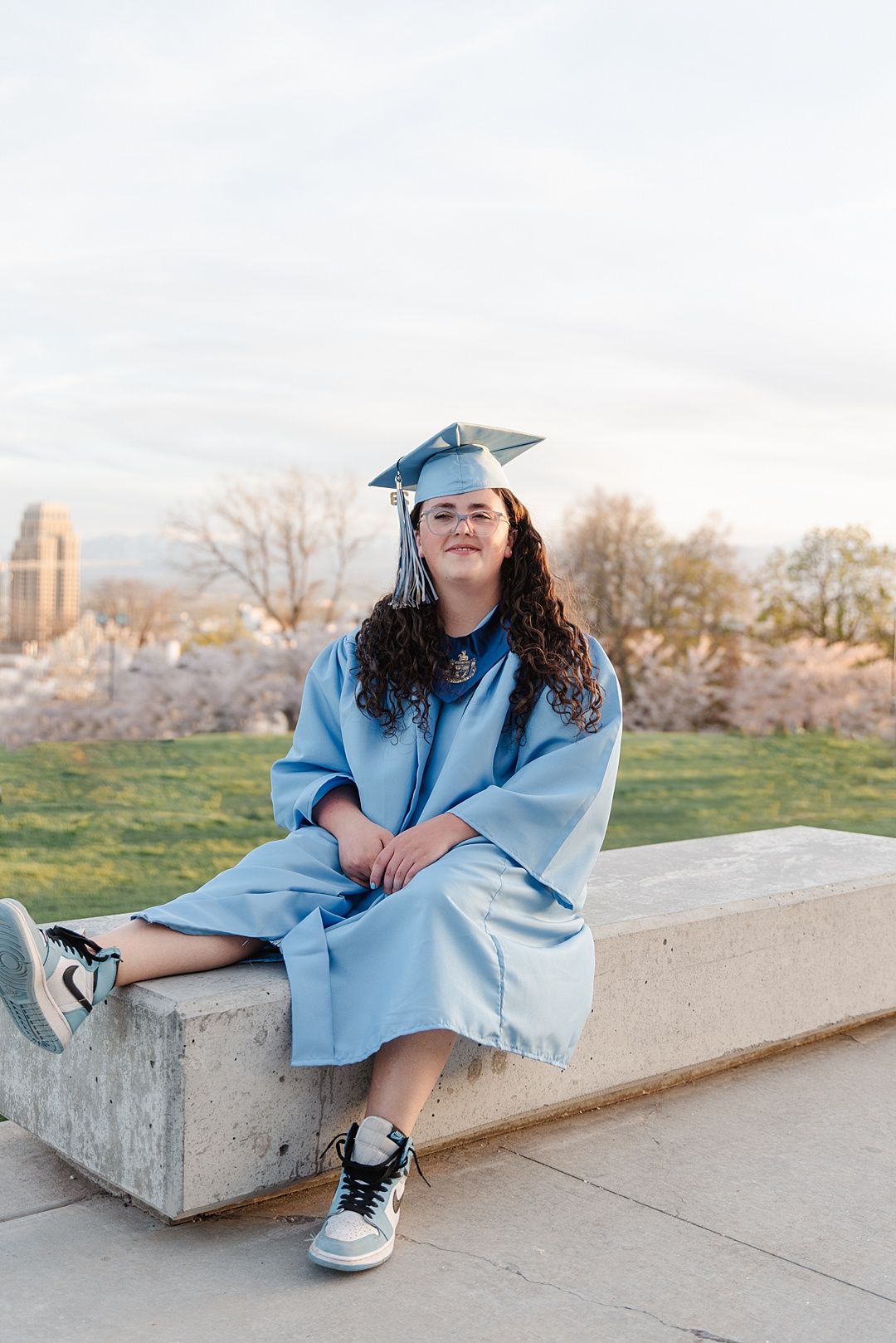 Graduate in light blue cap and gown sits on concrete ledge with cherry blossoms in background.